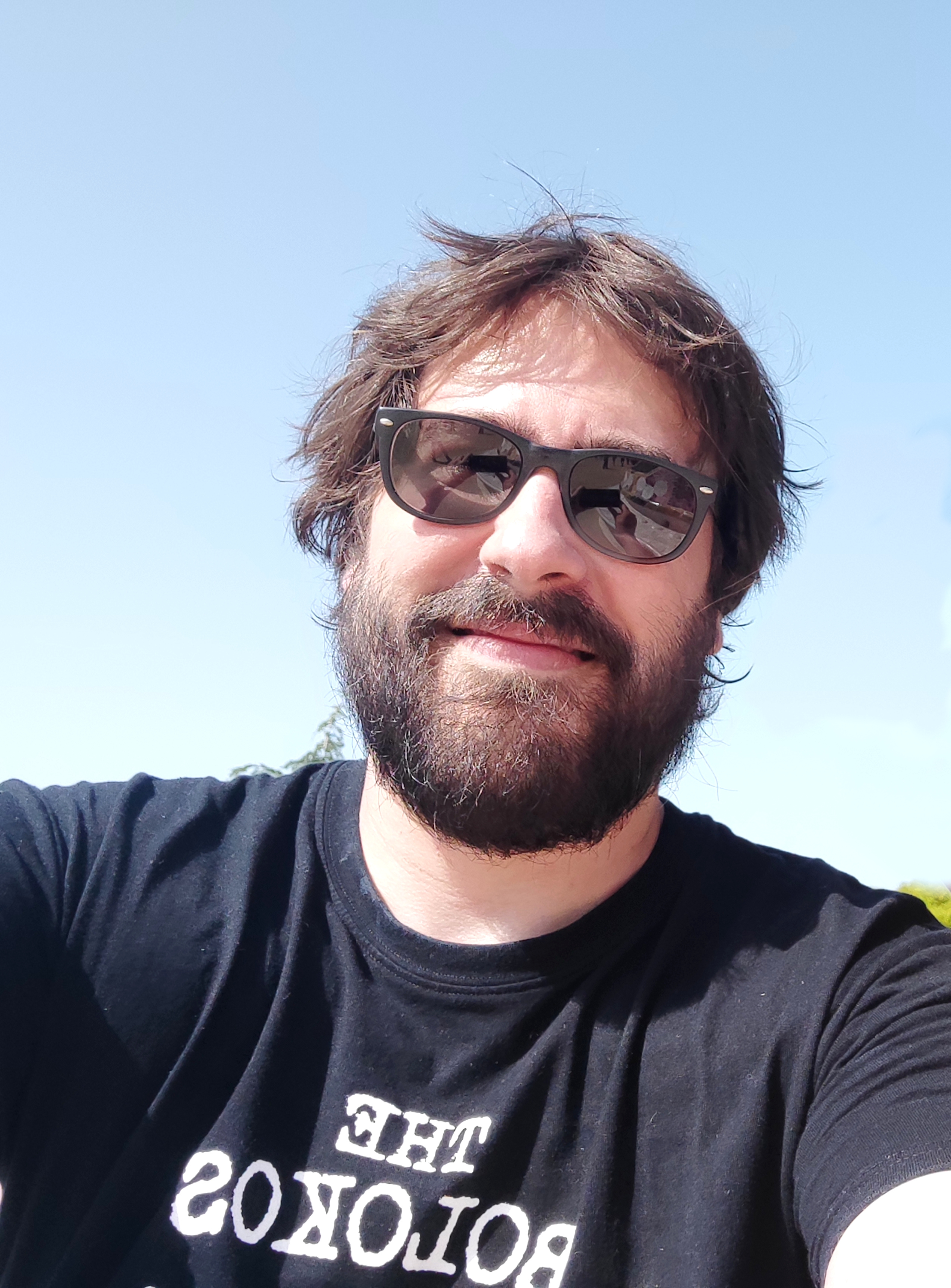 Nicolas souriant à la table d'un café. Il porte un T-shirt bleu foncé à l'éffigie de l'équipe de hockey Colorado Avalanches et est caché derrière une casquette noire, des lunettes de soleil noires et il tient une pinte de bière à la main droite